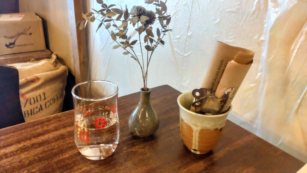 ネクタイコーヒーのテーブルに並ぶ、レトロな花柄のグラスとメニューを入れた陶器のカップ。一輪挿しのドライフラワーに細やかな気配りが感じられる。