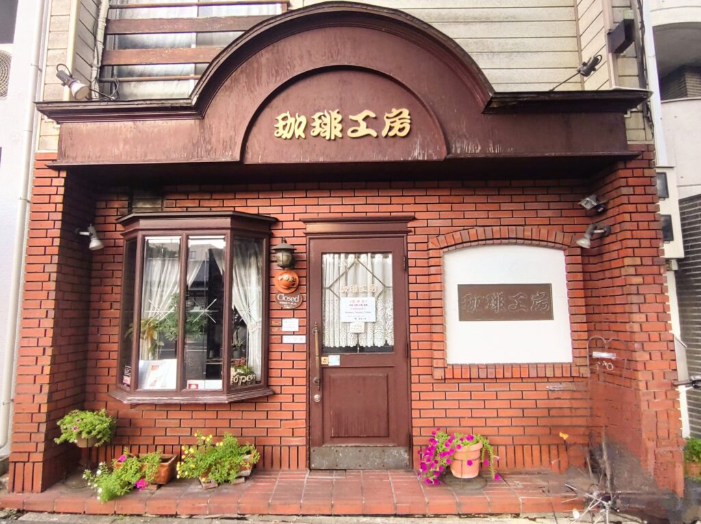 レンガ造りの外観が印象的な喫茶店「珈琲工房」の入口。木製ドア、窓辺に花が飾られ、昔ながらの温かみを感じる雰囲気